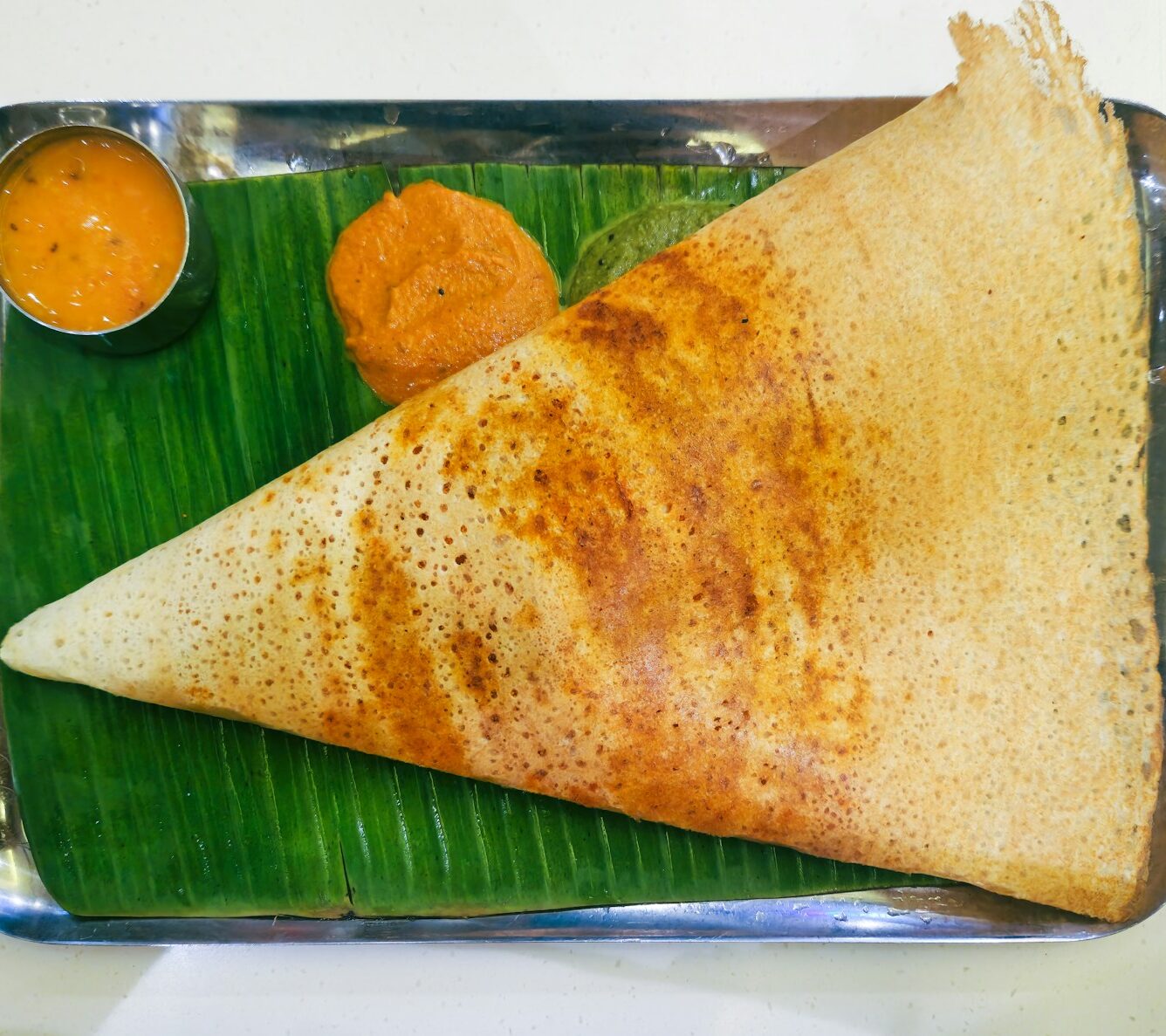 A crispy dosa with dips on a silver tray.